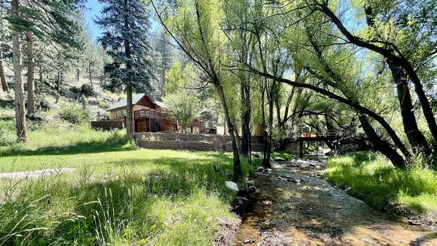 A rustic cabin with a stone wall and wooden deck, surrounded by lush greenery and a clear stream. - The Dam Overlook  • 3 Min Walk to the Lake • Hot Tub • Game Room (Evergreen)