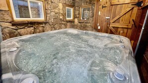 A hot tub in a rustic room with stone walls and wooden beams, featuring two windows and a wooden door. - The Dam Overlook  • 3 Min Walk to the Lake • Hot Tub • Game Room (Evergreen)