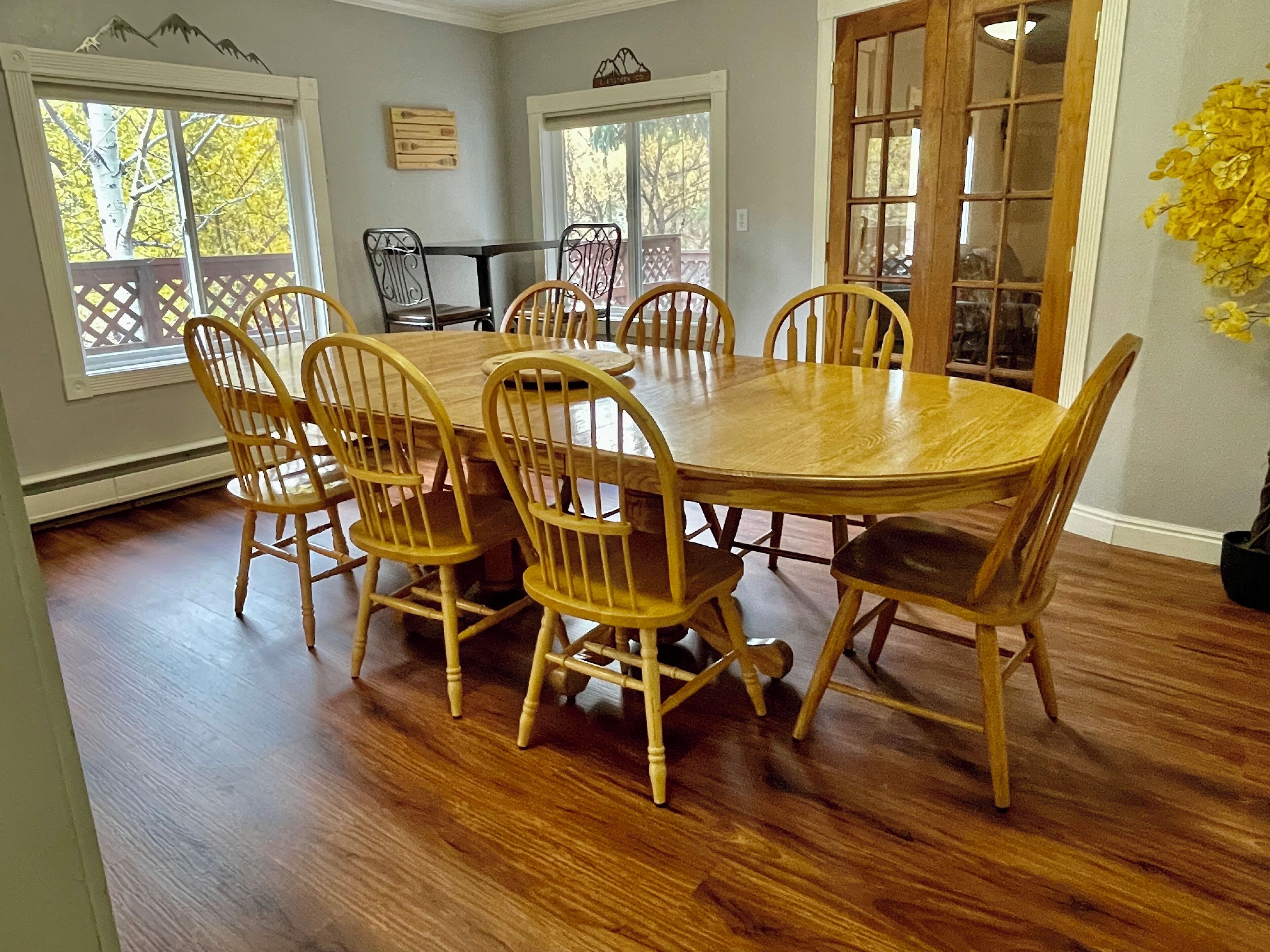Uma sala de jantar com uma grande mesa de madeira rodeada por seis cadeiras, janelas amplas com vista para árvores e piso de madeira.