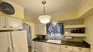 A kitchen with white cabinets, a black clock, a coffee maker, and a window with checkered curtains. - Country Club Charm (Omaha)