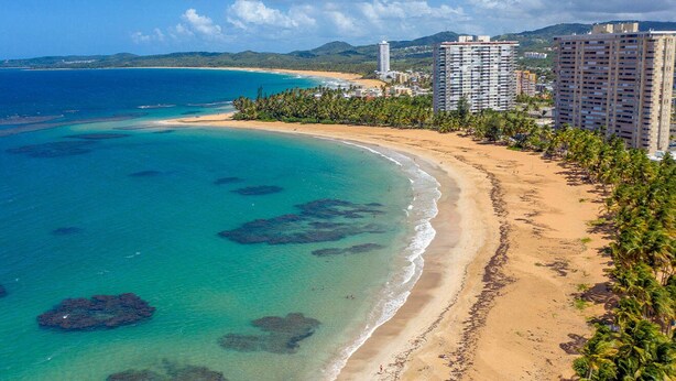 A coastal area with a beach, clear blue waters, and tall buildings in the background. - AQUA VILLA steps from the beach (Luquillo)