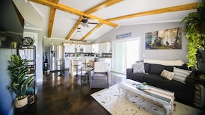 A spacious living area with a kitchen in the background, wooden beams on the ceiling, and a large window providing natural light. - Newly constructed cabin on a quiet residential pond.  (North Pole)
