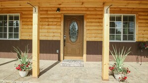 A wooden building with a central door, flanked by two windows, and two potted plants with red flowers. - Newly constructed cabin on a quiet residential pond.  (North Pole)