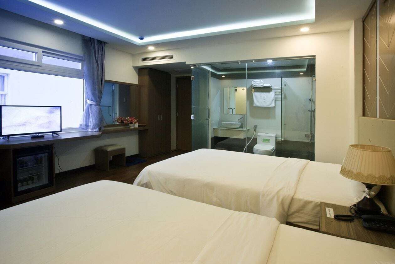 A hotel room with two beds, a television, a small table with a lamp, and a bathroom with a glass shower enclosure.