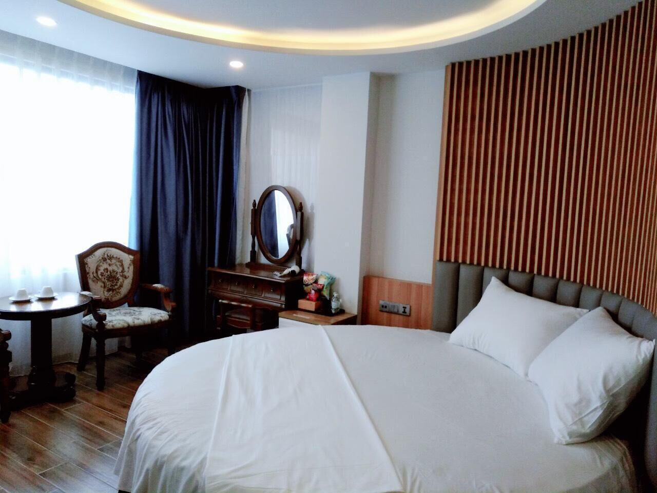 A hotel room with a large bed, a wooden headboard, a round mirror, a wooden dresser, a small table with cups, and a chair.