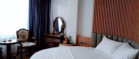 A hotel room with a large bed, a wooden headboard, a round mirror, a wooden dresser, a small table with cups, and a chair.