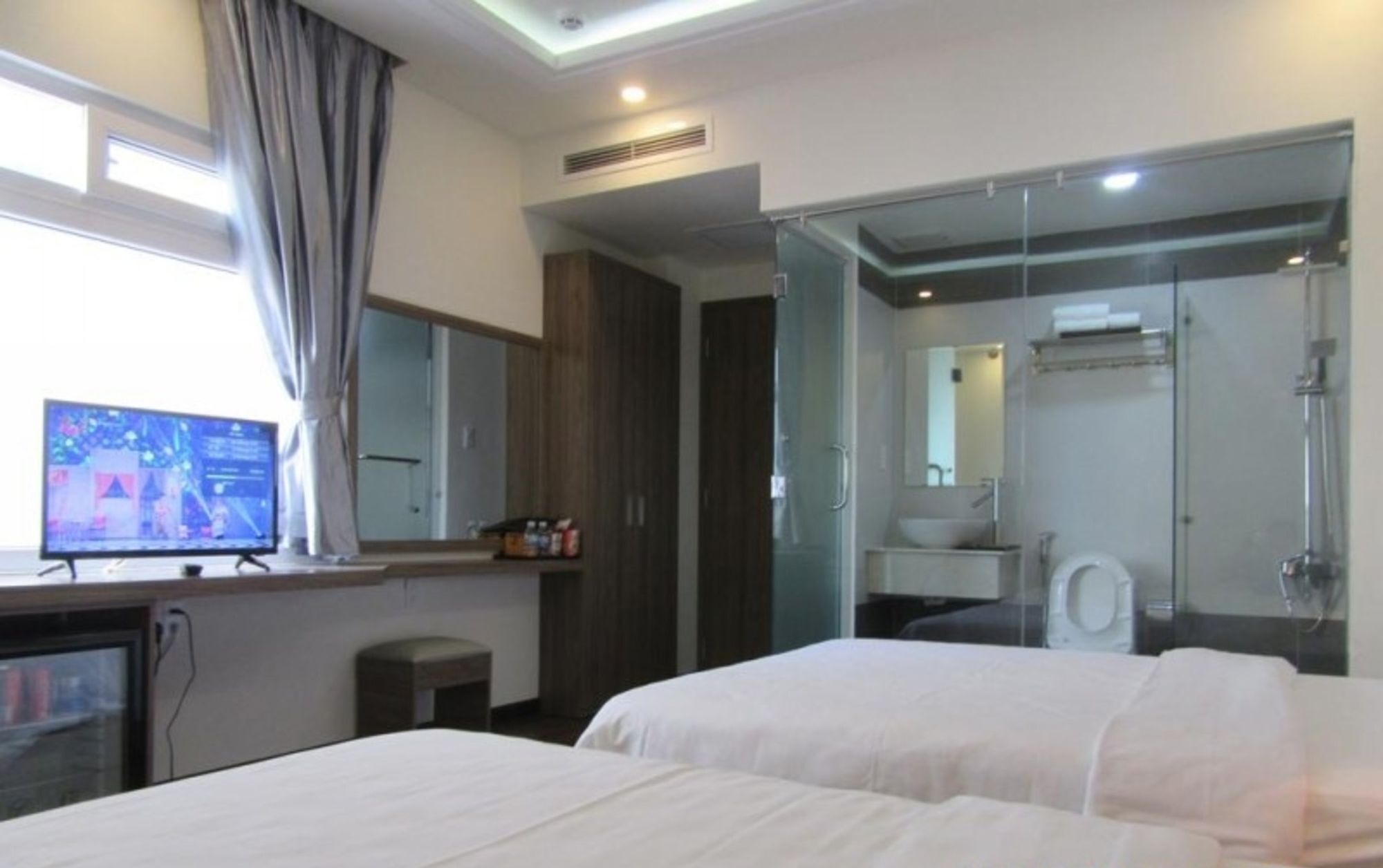 A hotel room with two beds, a TV, a desk, a bathroom with a glass shower enclosure, and a large mirror.