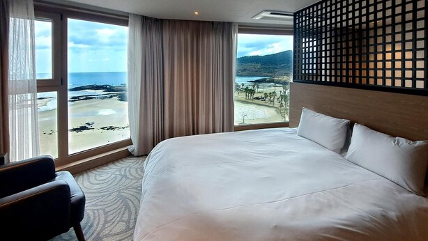 A hotel room with a large bed, a chair, and a view of a beach and mountains. - Saint Beach Hotel (Jeju City)