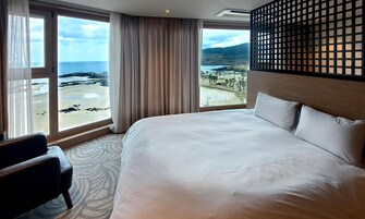 Habitación de hotel con una cama grande, una silla y vistas a la playa y las montañas.