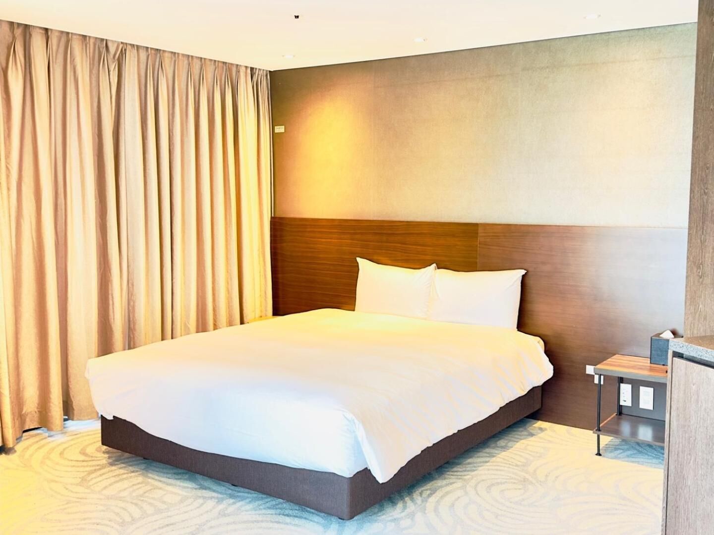 Habitación de hotel con una cama grande, un cabecero de madera, una mesita de noche y una cortina.
