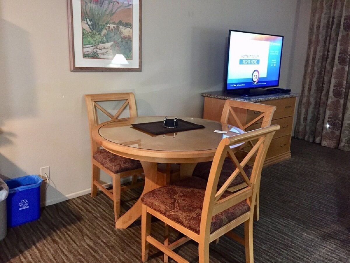 Ein Hotelzimmer mit einem runden Holztisch, vier Stühlen, einem Fernseher auf einem Schrank, einem gerahmten Bild an der Wand und einem blauen Mülleimer.