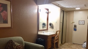 A compact hotel room with a green sofa, a wooden desk, a small bathroom with a mirror, and a door. - Studio in Palm Springs 2 (Palm Springs)