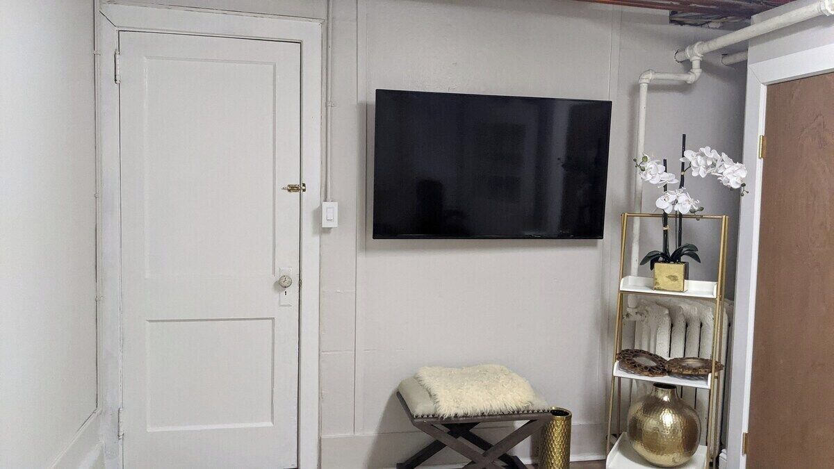 A room with a white door, a mounted flat-screen TV, a small cushioned stool, and a decorative shelf with a vase and orchids.