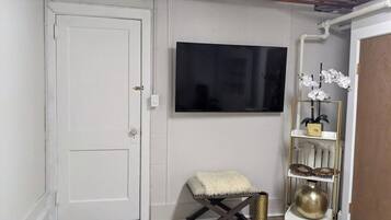 A room with a white door, a mounted flat-screen TV, a small cushioned stool, and a decorative shelf with a vase and orchids.
