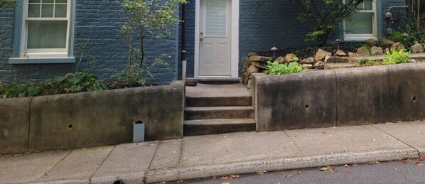 Un edificio residencial con una puerta blanca, una escalera de hormigón y un pequeño jardín con rocas y plantas.