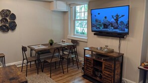 A dining area with a wooden table and chairs, a TV mounted on the wall, and a window with a view of trees.