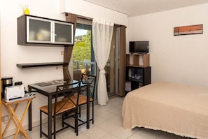 Una habitación con cama, televisión, mesa con sillas, una kitchenette pequeña y una ventana con cortinas.