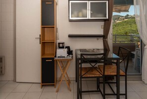 Eine kompakte Küche mit Mikrowelle, Toaster und Kaffeemaschine auf einem kleinen Tisch. Ein Holzkorpus mit offenen Regalen, ein schwarzer Stuhl und ein Glastisch sind zu sehen. Ein Fenster bietet Blick auf Grünfläche.