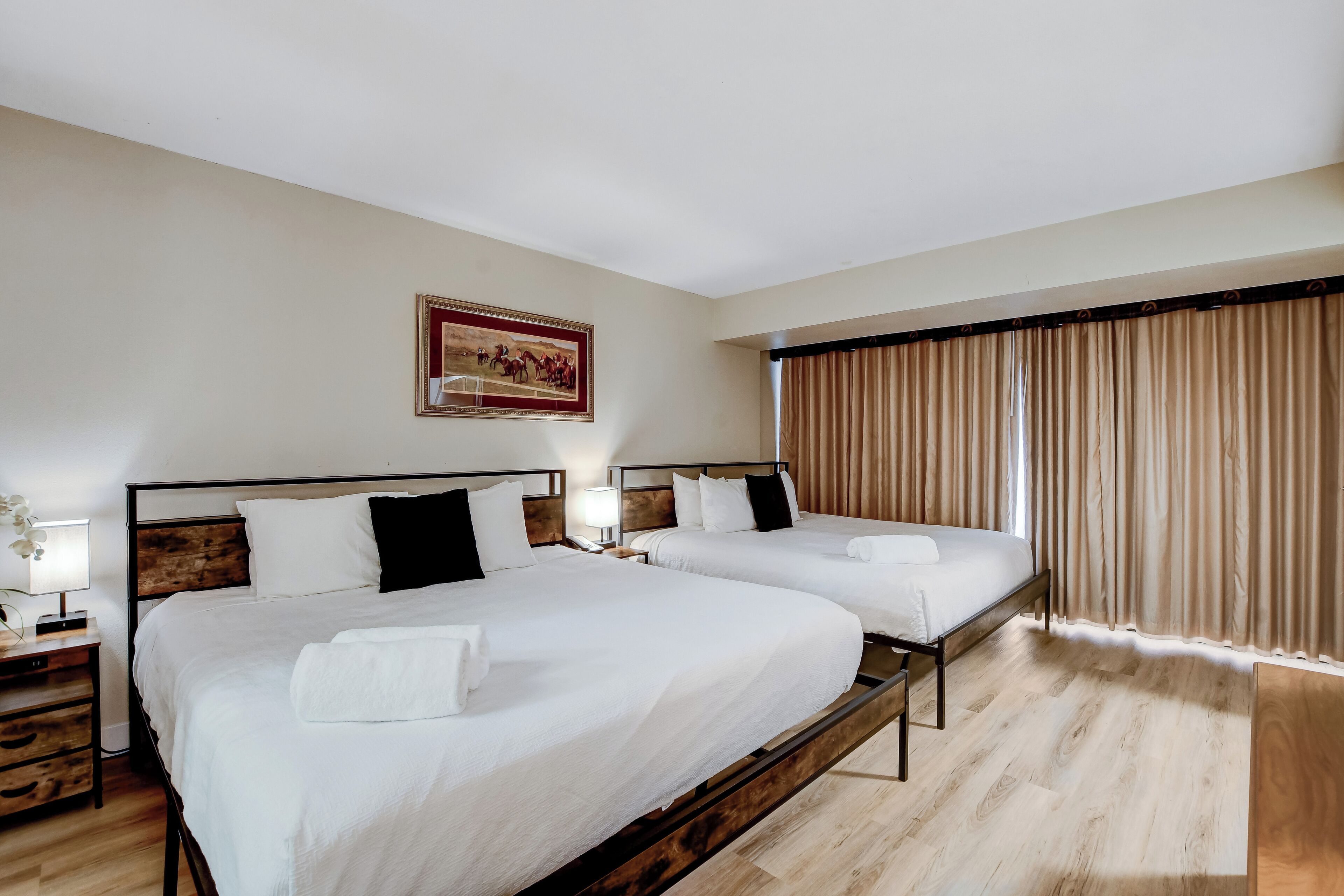 A hotel room with two beds, wooden headboards, a nightstand with a lamp, and a large window with curtains.