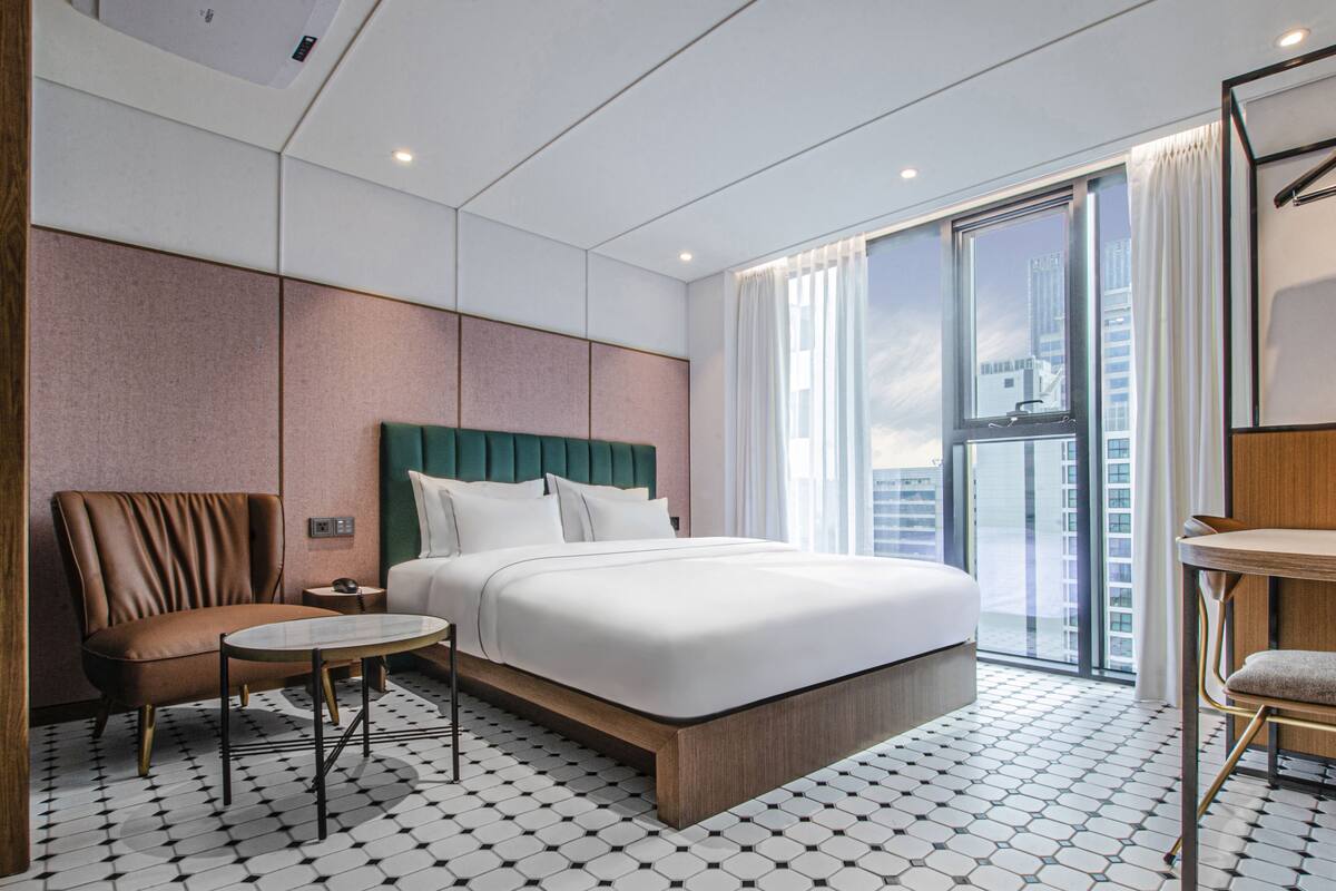 Une chambre d’hôtel moderne dotée d’un grand lit, d’un coin salon avec une table et offrant une vue sur la ville grâce à de grandes fenêtres.