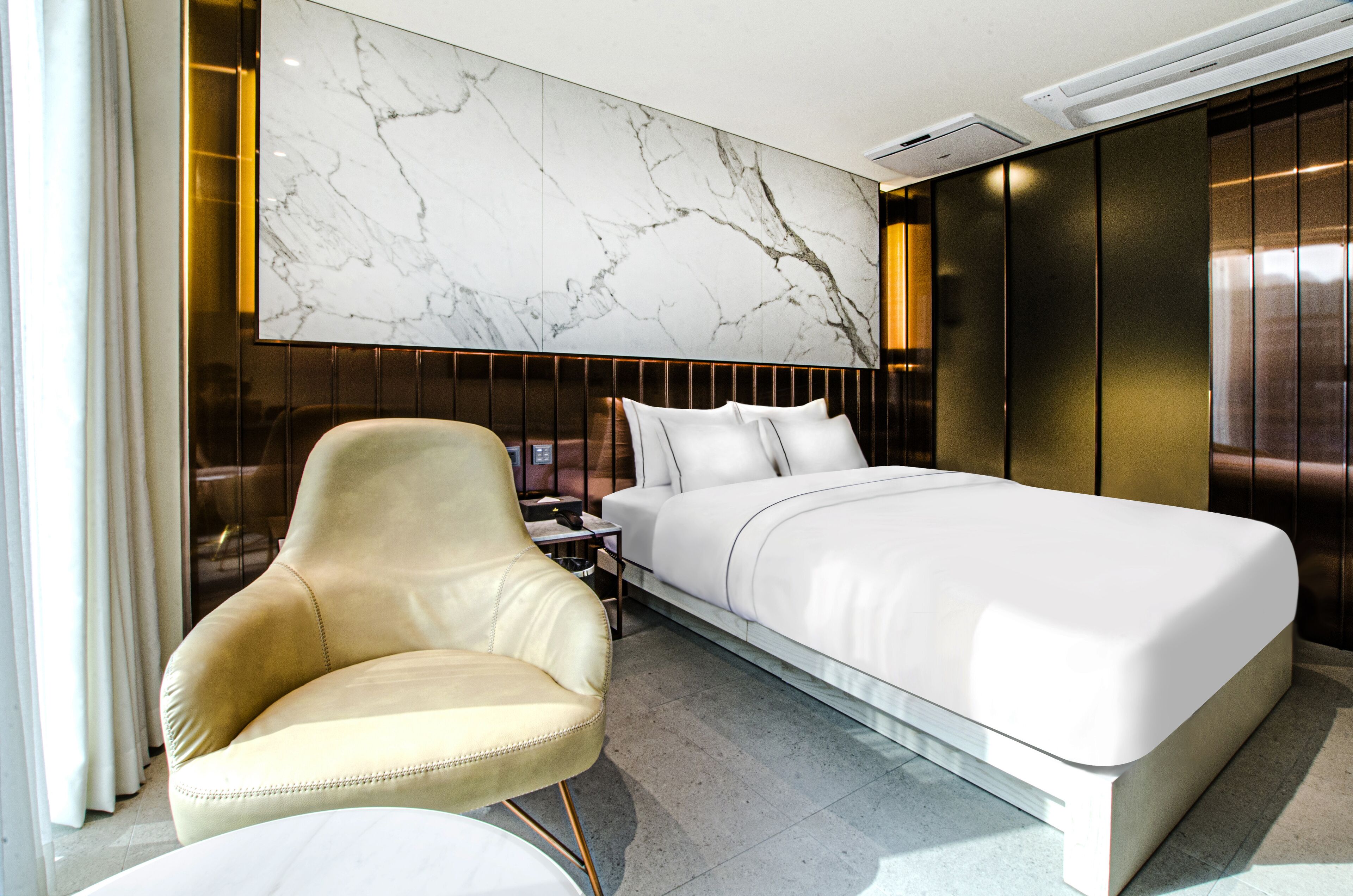 A modern hotel room with a large bed, a leather armchair, a marble wall, and a large window.