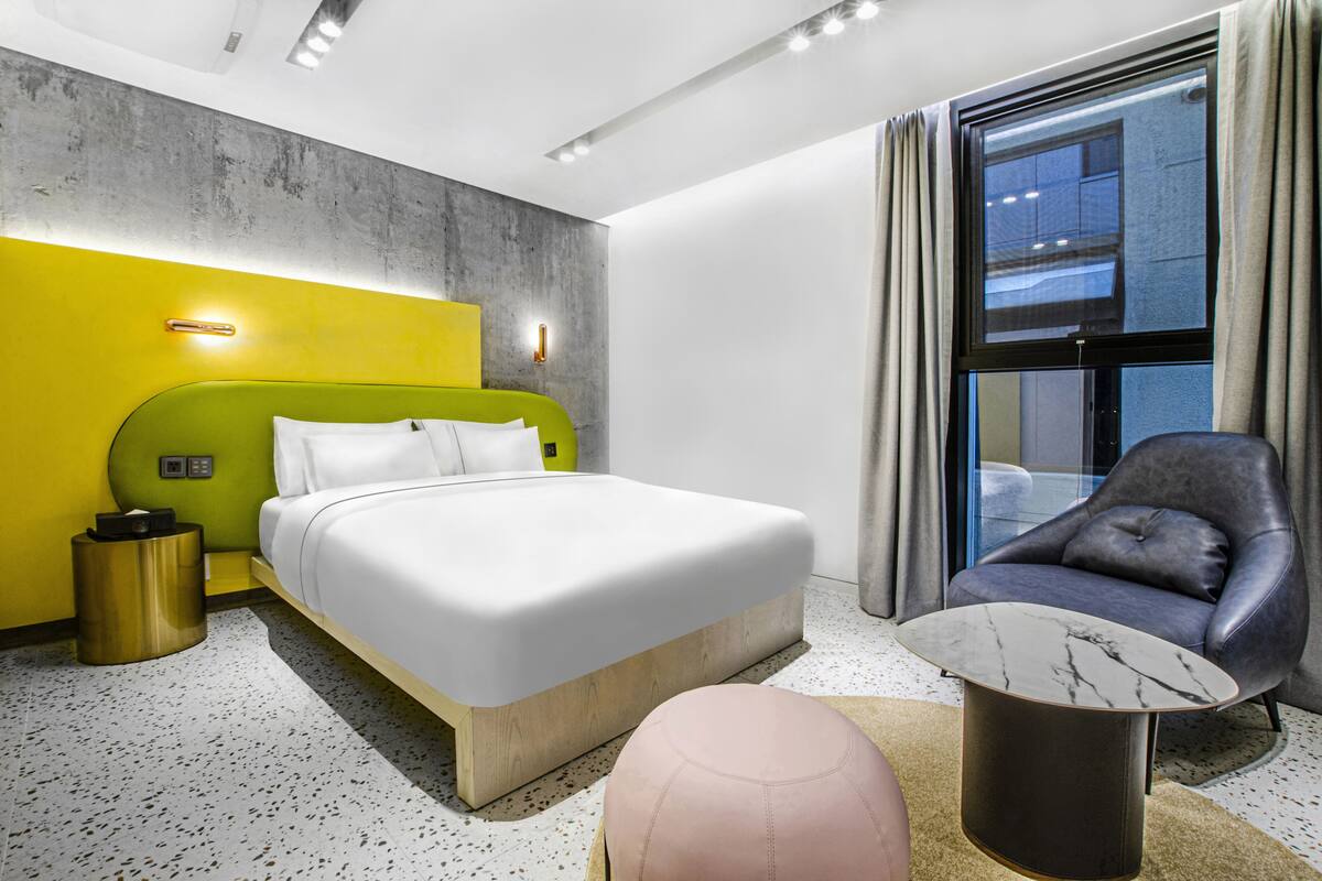 Une chambre d’hôtel moderne avec un mur d’accent vert, un grand lit, un fauteuil en cuir et une table basse en marbre.