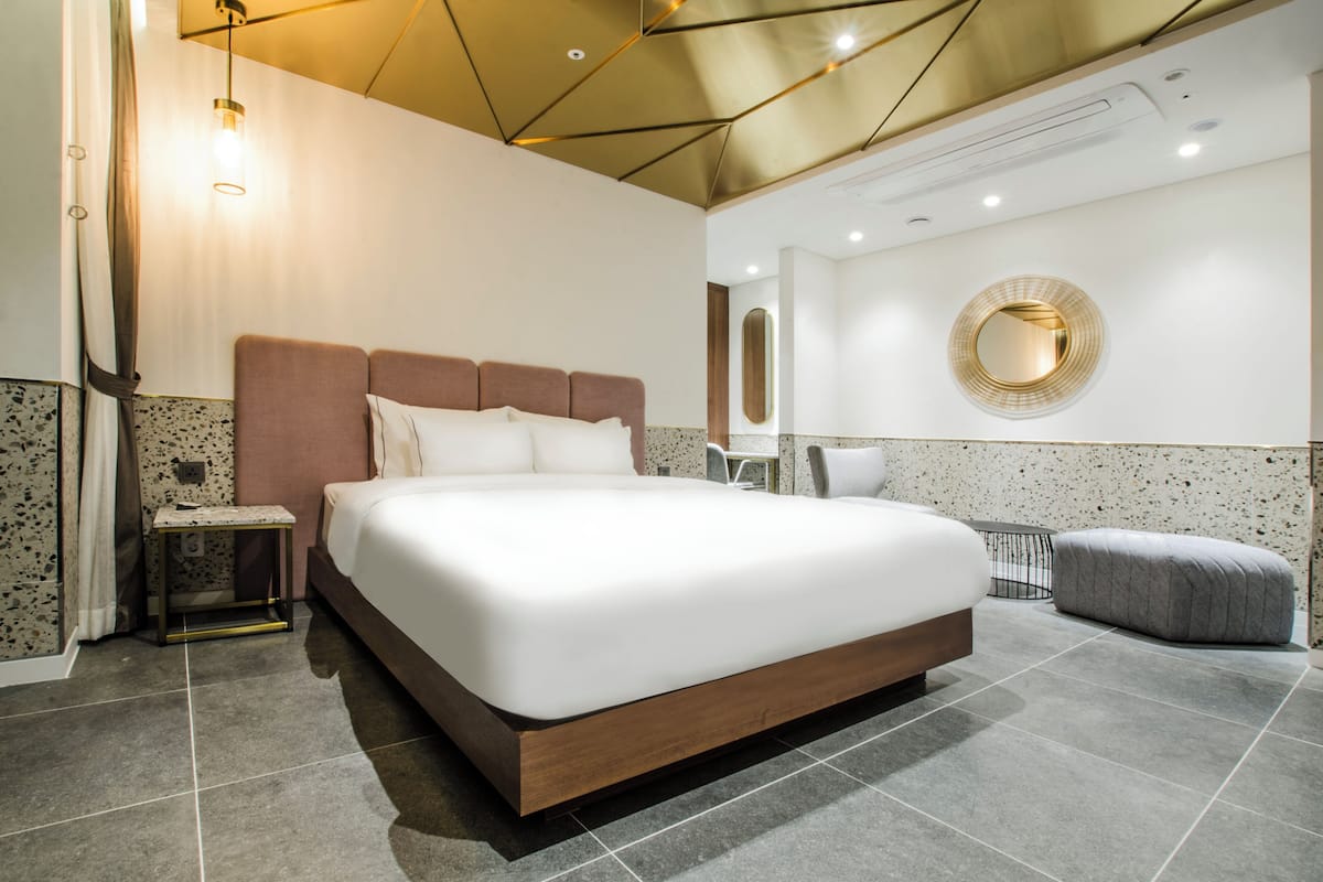 Une chambre d’hôtel moderne avec un grand lit, une table de chevet, un miroir rond et un pouf gris.