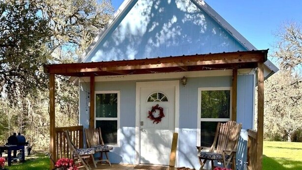 Een klein, gelijkvloers huis met een veranda, een krans op de deur en twee stoelen op de veranda.