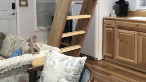 Een kamer met een houten trap naar een slaapverdieping, een blauwe stoel met een gestreept kussen, een bank met bloemenprint en houten kasten.