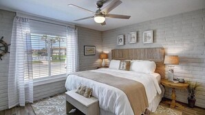 Un dormitorio con cama, ventilador de techo, ventana con cortinas, mesita de noche con lámpara y un reloj en la pared.