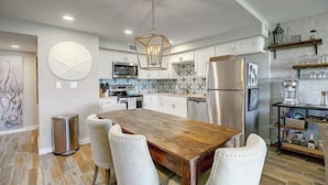 Una cocina moderna con una mesa de comedor de madera, sillas blancas, electrodomésticos de acero inoxidable y una lámpara colgante.