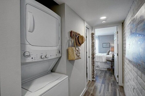 Un cuarto de lavandería con lavadora y secadora integradas, un pasillo que conduce a un dormitorio con cama y mesita de noche, y una pared con diseño texturizado.