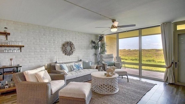 Un salón espacioso con pared de ladrillo blanco, mobiliario de mimbre, un ventanal con vistas al exterior y un ventilador de techo.