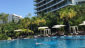 En swimmingpool med liggestole og parasoller, omgivet af palmer og en hotelbygning i flere etager.