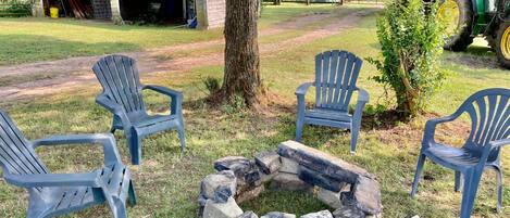 A rural outdoor setting with a fire pit, two Adirondack chairs, and a tractor in the background.
