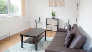 A modern living room with a dark brown sofa, a glass coffee table, and a small side table with plants. - Large Shoreditch, Hoxton 3-Bed Garden Flat (London)
