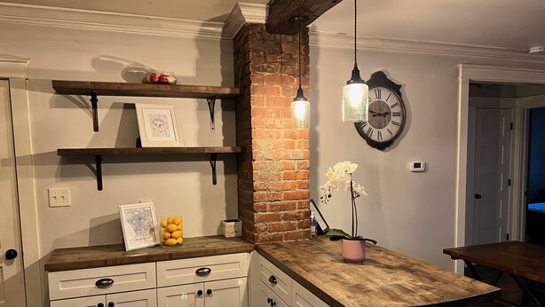 A kitchen with white cabinets, a brick accent wall, pendant lighting, and a clock on the wall. - Very Welcoming (East Stroudsburg)