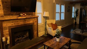 A living room with a fireplace, a television mounted above it, a sofa, a coffee table, and a potted plant. - Very Welcoming (East Stroudsburg)