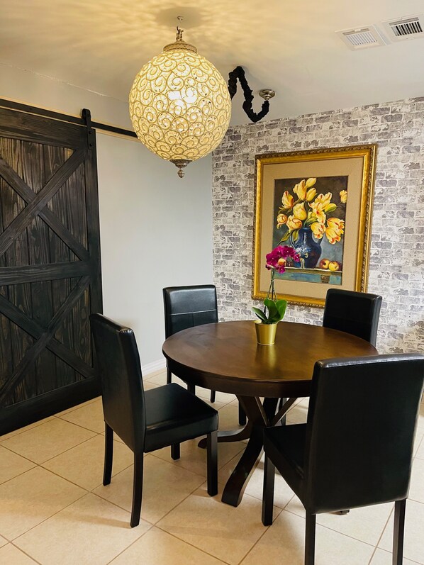 A dining area with a round table, black chairs, a decorative hanging light fixture, a framed painting of flowers, and a brick wall.