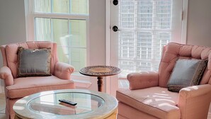A room with two pink armchairs, one with a patterned cushion and the other with a solid cushion, a glass coffee table with a remote control, and a small wooden table with a decorative top. - Momo's sweet home 2BR/2BA (Dallas)