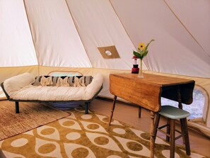 Ein Zeltinnenraum mit einem Bett, einem Holztisch, einem Stuhl und einer kleinen Tischlampe.