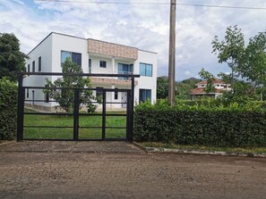 A modern two-story building with a white facade and a brown roof, surrounded by greenery and a paved area in front.