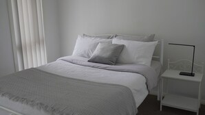 Een net opgemaakt bed met wit beddengoed en een grijze plaid, een witte nachtkastje met een afstandsbediening, en een raam met verticale jaloezieën.