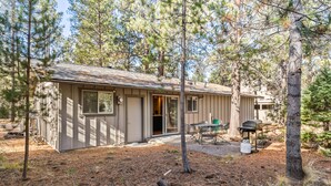 A small cabin with a front door, a patio with a table and chairs, and a grill area. - Walk to Village, Sharc Passes, Air Conditioning, King Bed, Bikes (Sunriver)