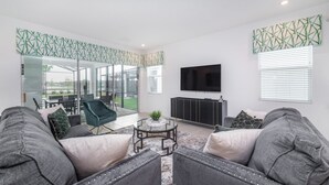 A modern living room with grey sofas, a glass coffee table, and a flat-screen TV mounted on the wall.