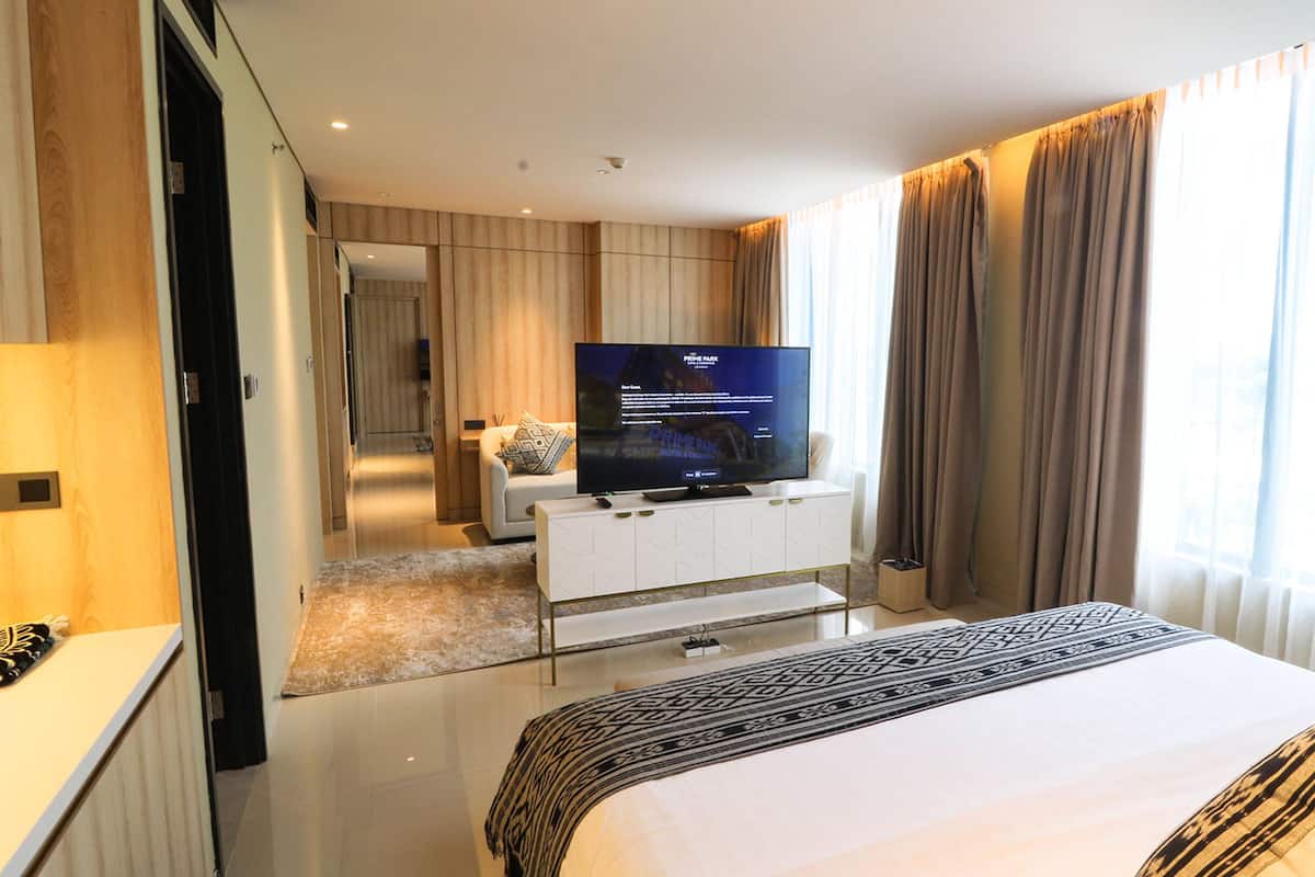 A modern hotel room with a large bed, a flat-screen TV on a white console, a walk-in closet, and a view of the city through large windows.