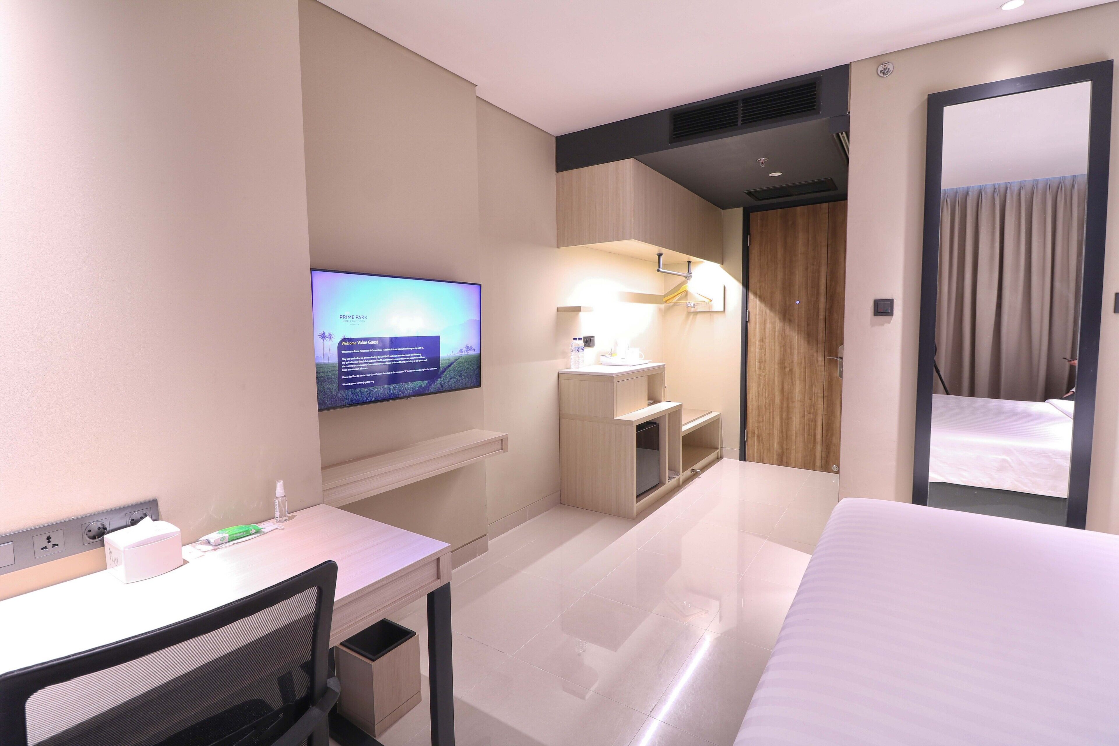 A hotel room with a bed, a desk with a computer, a TV, a mirror, and a bathroom with a shower.