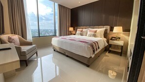 A modern hotel room with a large bed, a sitting area with a chair, a bedside table with a lamp, and a view of the city through large windows. - Prime Park Hotel & Convention Lombok (Mataram)