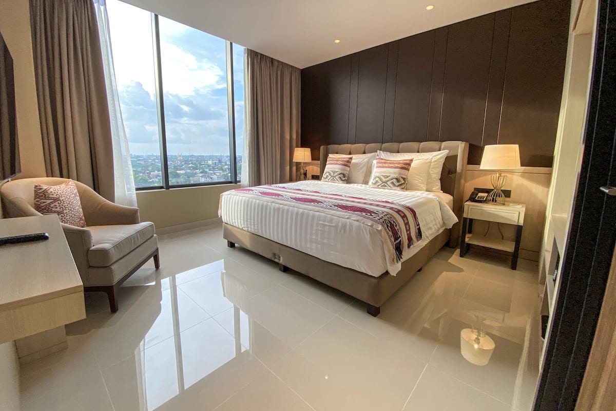 A modern hotel room with a large bed, a sitting area with a chair, a bedside table with a lamp, and a view of the city through large windows.