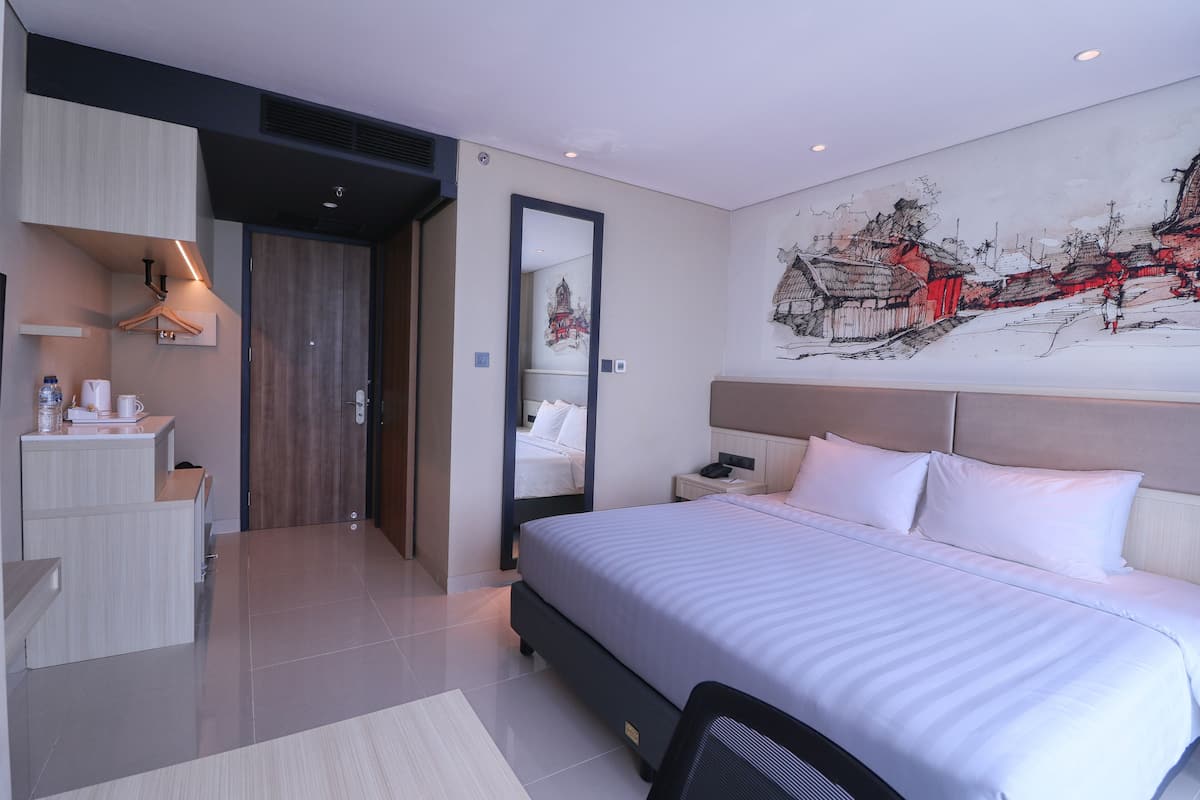 A modern hotel room with a large bed, a desk with a chair, a wardrobe, and a wall-mounted artwork.